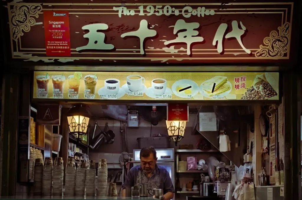 A detailed interior view of 'The 1950's Coffee' (五十年代) hawker stall in Singapore. The vendor is busy behind the counter, surrounded by stacks of paper cups and traditional porcelain cups. The stall features hanging lantern lights, a menu with prices for various coffees and teas, and a Michelin-recommended sticker for 2016This is a quintessential traditional coffee stall experience in a Singapore hawker center.