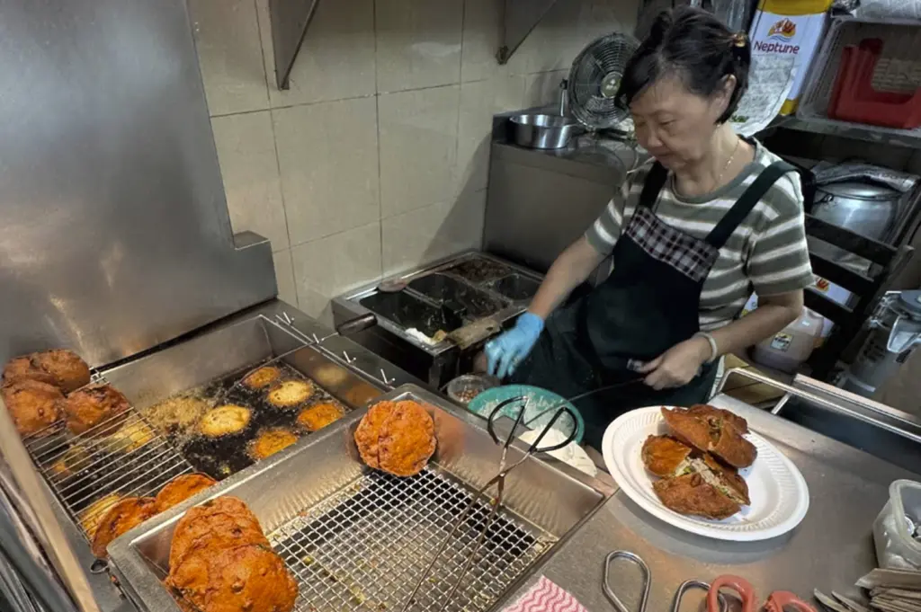 A high-angle, close-up shot inside a hawker stall focusing on the preparation of Fuzhou Oyster Cakes. An elderly woman in a striped shirt and dark apron works behind a stainless steel counter, wearing blue latex gloves. To the left, several golden-brown, disc-shaped oyster cakes drain on a wire rack above a deep fryer filled with bubbling oil. On the right, a white plate holds two freshly fried, halved oyster cakes, revealing a savory filling of greens and meat.