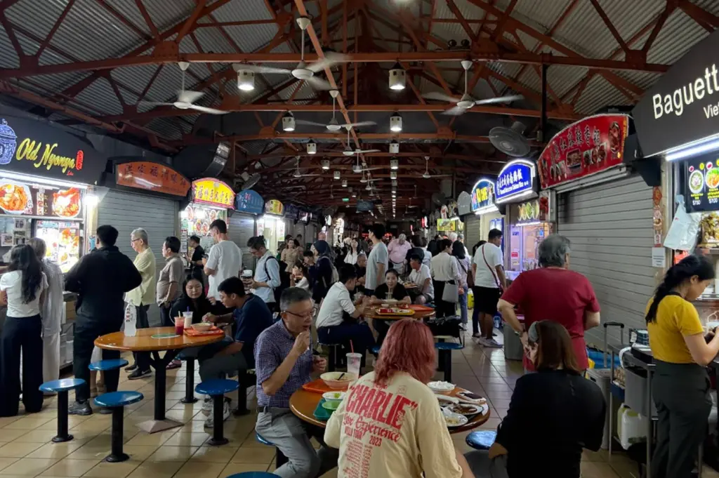 A wide shot of a bustling, open-air hawker centre with a high, red-beamed industrial ceiling and numerous ceiling fans. Various food stalls like "Old Nyonya" and "Baguette" line the sides. The center is filled with people sitting at round tables and walking through the aisles, creating a vibrant, casual dining atmosphere.