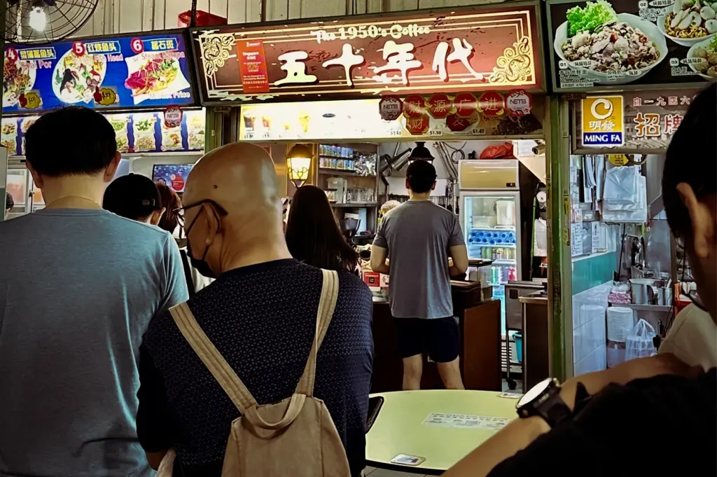 A busy morning queue of customers waiting at The 1950's Coffee (五十年代) hawker stall in. A traditional Nanyang kopi vendor in a Singapore hawker center, featuring authentic signage, vintage lanterns, and a bustling food court atmosphere.