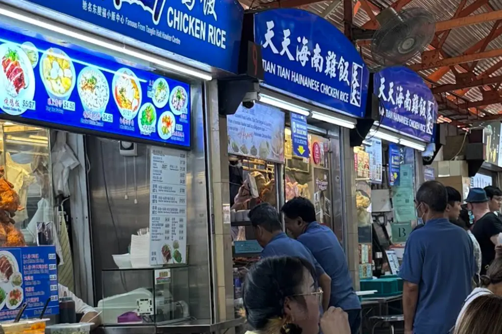 A medium shot featuring the iconic blue neon signage of "Tian Tian Hainanese Chicken Rice" at stalls 10 and 11. The vibrant blue signs stand out against the industrial ceiling. Below the signs, a menu board displays various chicken rice sets with prices. A few customers are seen at the counter interacting with the staff. To the left, another stall, "Ah Tai Hainanese Chicken Rice," is partially visible with its own illuminated menu of roasted meats.