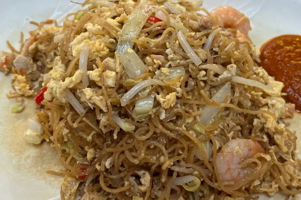A close-up, top-down view of a plate of Sin Chow Bee Hoon (Singapore-style fried vermicelli). The thin rice noodles are stir-fried to a golden-brown color and mixed with scrambled eggs, bean sprouts, small pieces of pork, and large, pink shrimp. A dollop of bright red sambal chili paste is served on the side.