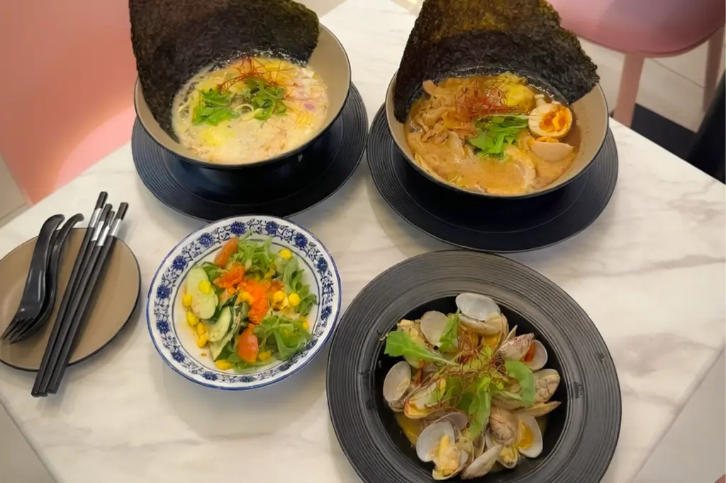 A delicious Japanese meal spread from Ramen Story, featuring two steaming bowls of ramen topped with large sheets of nori and soft-boiled eggs, a side of fresh garden salad with corn and cherry tomatoes, and a flavorful plate of stir-fried clams garnished with greens.