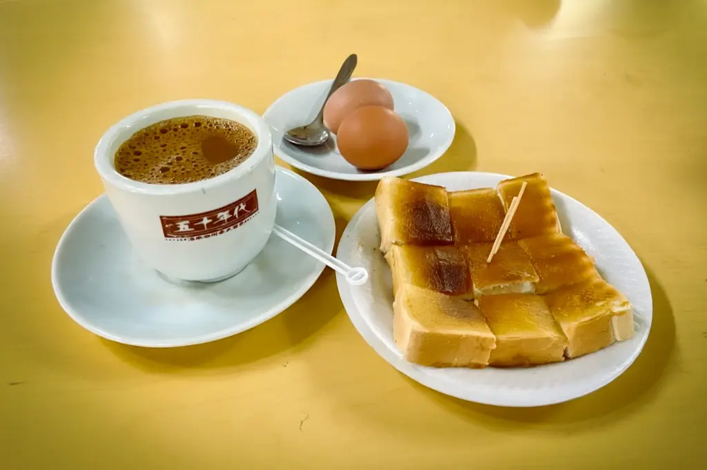 A close-up shot of a traditional Singaporean Nanyang breakfast from "The 1950s Coffee" stall, featuring kaya butter toast cut into squares, two soft-boiled eggs, and a cup of frothy hot coffee with the stall's logo on a yellow table.