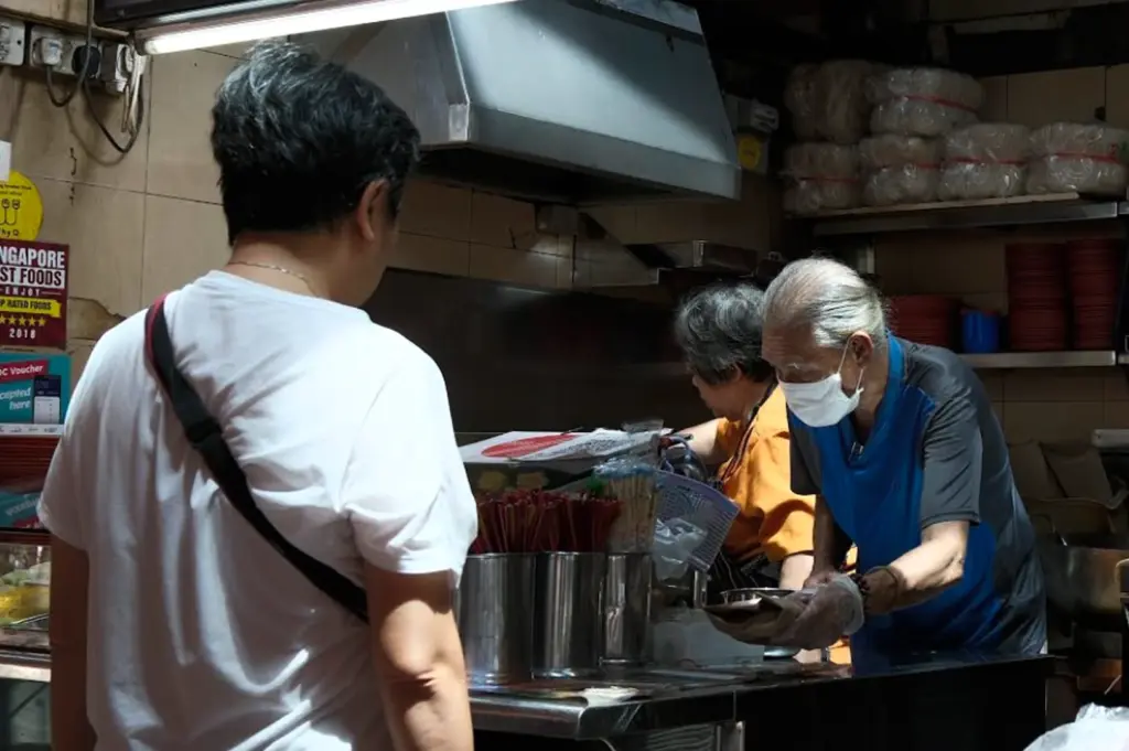 A candid, low-light shot from behind a customer in a white t-shirt looking into a hawker stall. Inside the compact, stainless steel kitchen, an elderly man wearing a face mask and blue shirt leans forward to hand over a wrapped food item. In the background, a woman in an orange shirt works near a large exhaust hood. Stacks of clear plastic-wrapped noodle bundles sit on a shelf above the prep area, and a "Singapore Best Foods" award sticker is visible on the wall.