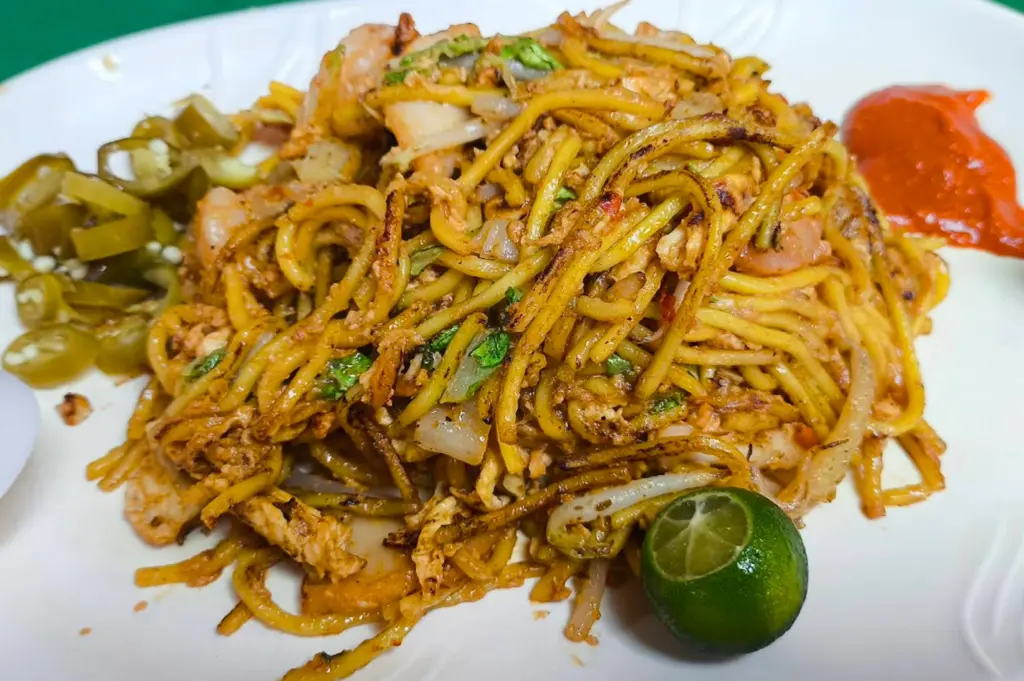 A vibrant plate of Mee Goreng featuring thick, yellow egg noodles stir-fried with a reddish-orange spice paste. The dish includes charred bits, green leafy vegetables, bean sprouts, and shrimp. It is garnished with a small green calamansi lime and served with a side of sliced pickled green chilies and a splash of red chili sauce.