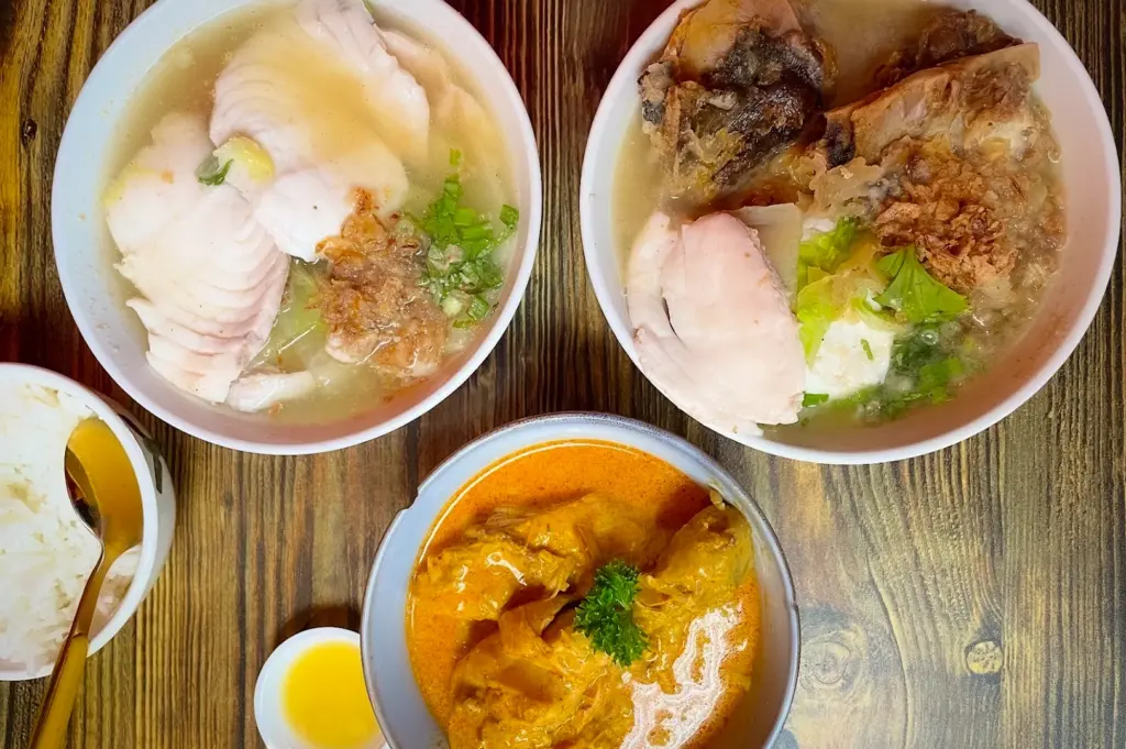 A flavorful traditional spread from Diners' Shack, featuring two bowls of sliced fish soup garnished with fried shallots and greens, alongside a rich, aromatic bowl of chicken curry, served with steamed white rice.