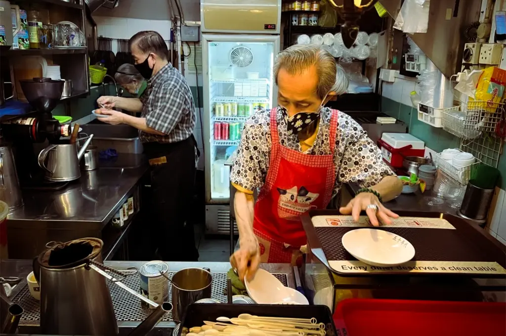 Inside the bustling "The 1950s Coffee" stall in Singapore, three elderly workers wearing face masks prepare food and drinks in a compact, traditional kitchen. In the foreground, a man in a red patterned apron and floral shirt arranges white bowls on a tray. To the left, another man in a plaid shirt handles soft-boiled eggs, while a third worker is partially visible in the background. The kitchen is packed with authentic details: a tall metal coffee pot with a cloth strainer, cans of condensed milk, stacks of plastic cups, and a refrigerator filled with colorful canned drinks. A sign on a tray reads, "Every diner deserves a clean table."
