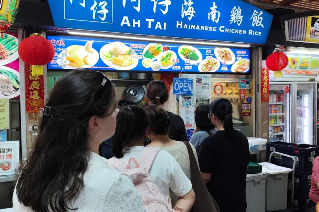 A eye-level shot from the back of a queue of people waiting at the "Ah Tai Hainanese Chicken Rice" stall. The focus is on the back of a woman with long, dark wavy hair in a white textured sweater. The stall's bright blue signage and a menu featuring photos of chicken rice, bean sprouts, and oyster sauce vegetables dominate the upper frame. Two red lanterns hang on either side of the stall, and a small "YES... We are OPEN" sign is hung near the service window.