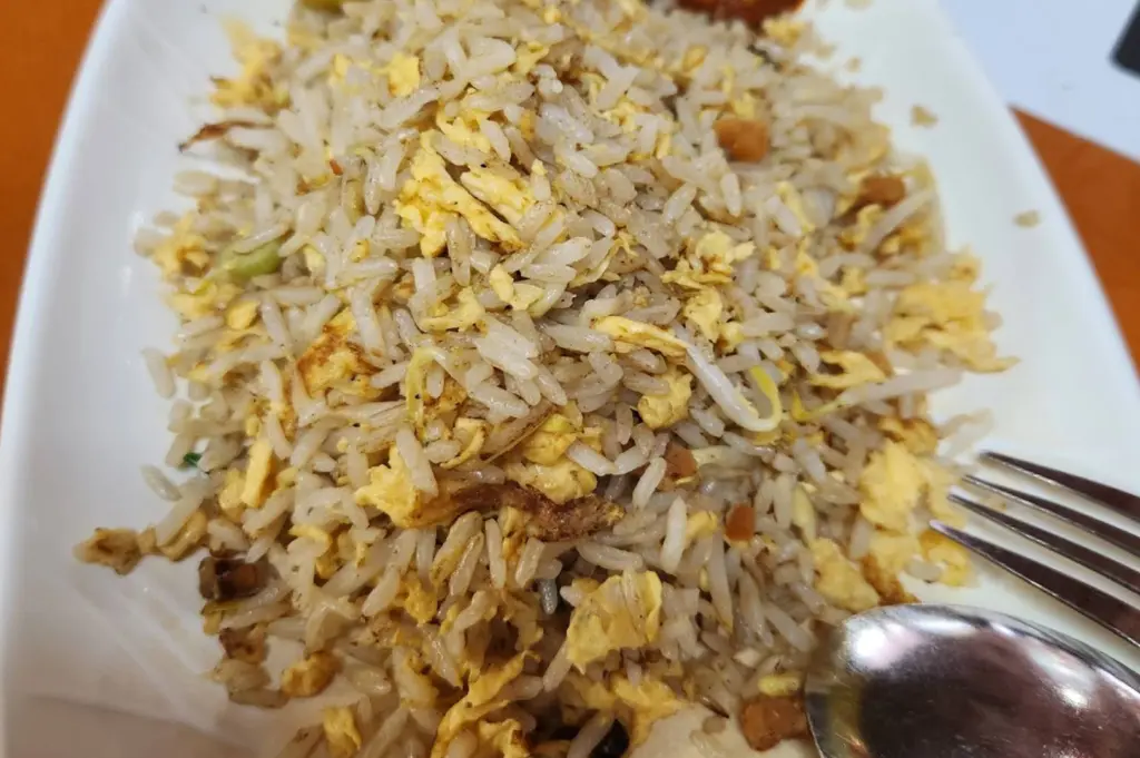 A close-up of a plate of Yangzhou Fried Rice. The white rice is fluffy and well-separated, tossed with generous amounts of scrambled egg, small cubes of savory char siew (BBQ pork), bean sprouts, and scallions. A metal spoon and fork rest on the side of the white patterned plate.