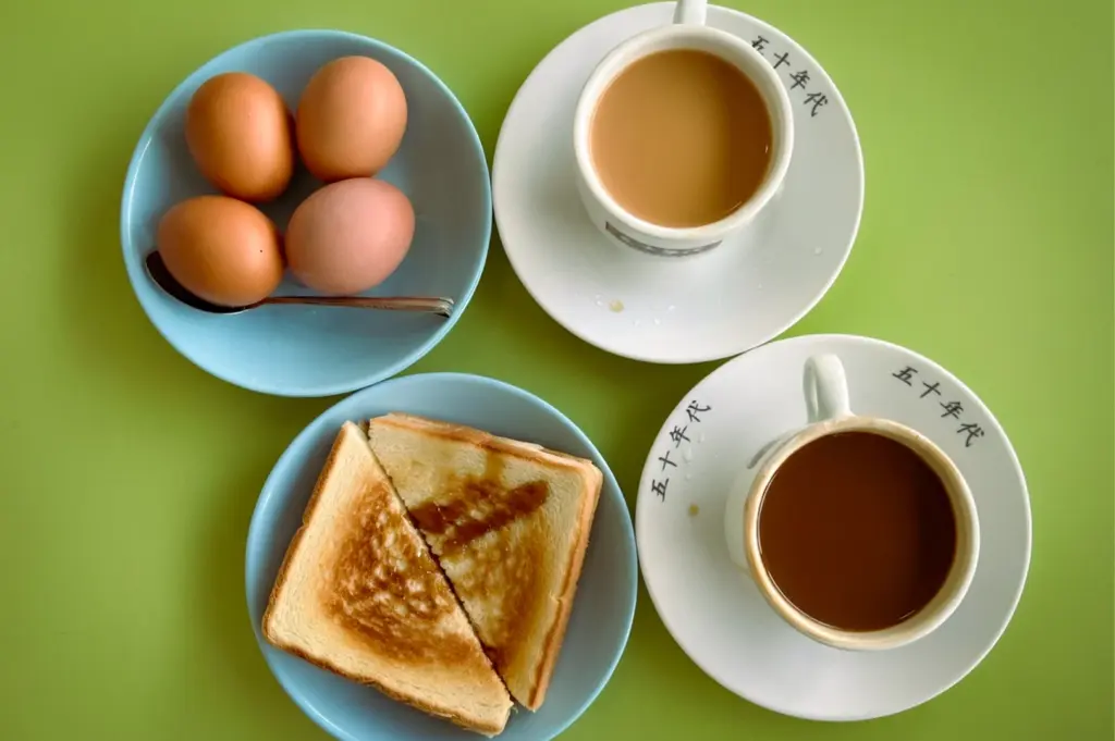 Top-down view of a traditional Singaporean breakfast at The 1950s Coffee. Two cups of Nanyang coffee, toast cut into triangles, and soft-boiled eggs in a blue bowl on a bright lime-green table.