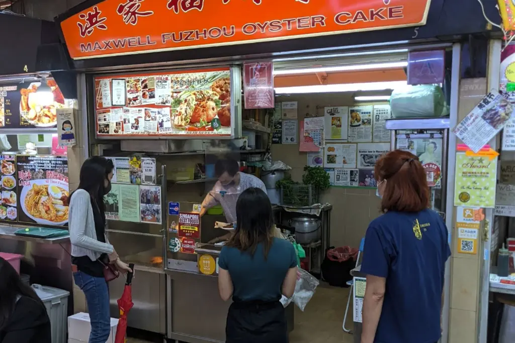 A straight-on shot of the "Maxwell Fuzhou Oyster Cake" stall (Stall 5). The stall features a large orange sign with white and black lettering. The front of the stall is covered in newspaper clippings, awards, and "Singapore Best Foods" stickers. A man in a grey shirt is visible inside the stall preparing food behind a clear plastic sneeze guard. Three customers stand in front of the counter, including a woman in a green shirt and another in a blue t-shirt.