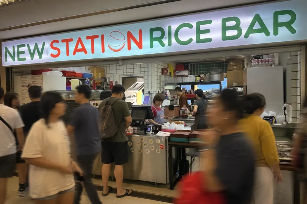 Busy New Station Rice Bar storefront with customers ordering at a casual eatery, featuring a bright sign, open kitchen, and fast-paced crowd in a modern food court setting