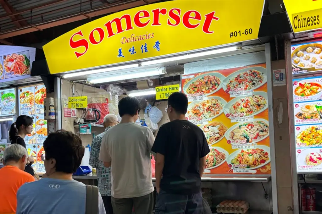 A clear view of the Somerset Delicacies stall (#01-60) during the day. Several customers are standing in line to order. The large pictorial menu prominently features eight different dishes, including Seafood Fried Rice, Beef Fried Rice, and Mixed Seafood Hor Fun, with prices listed between $4.00 and $5.50. Boxes of eggs are stacked at the base of the counter.