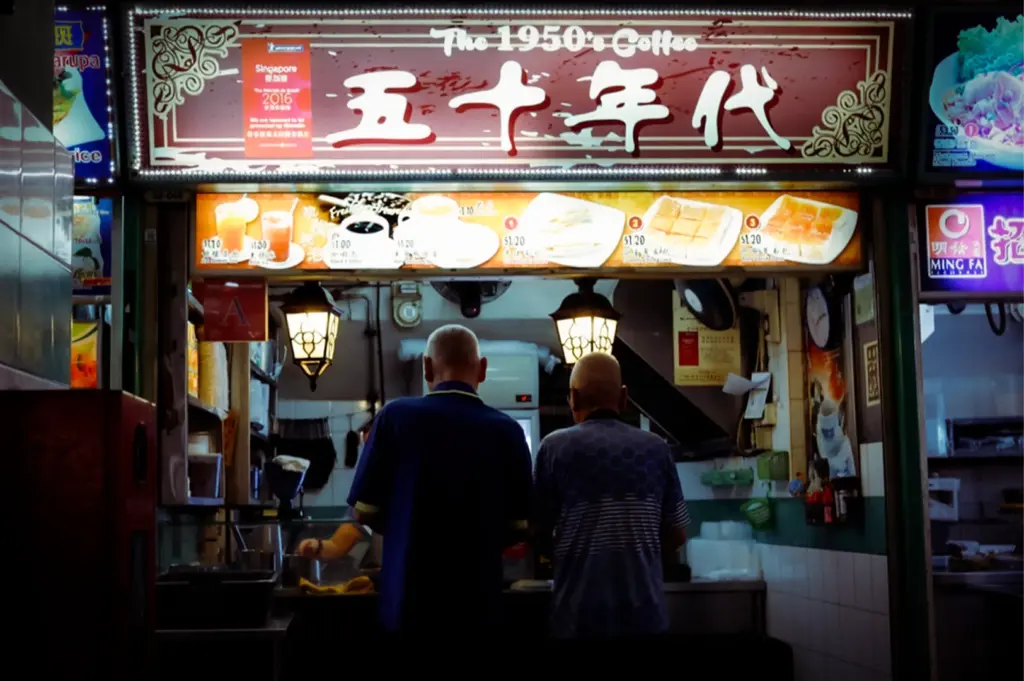Front view of The 1950s Coffee stall in Singapore, with two customers standing at the counter ordering kaya toast and soft boiled eggs beneath bright vintage signage, capturing a traditional kopitiam breakfast scene