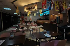A cozy Japanese-style ramen restaurant interior with dim ambient lighting, small square tables and pink chairs arranged throughout the dining area, hanging Japanese banners and lanterns decorating the ceiling, and a warmly lit service counter with digital menu screens in the background. The atmosphere feels intimate and modern, with dark wood furniture contrasting against soft lighting and colorful signage.