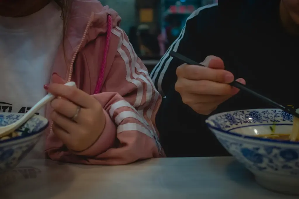 Two people eating noodle soup with chopsticks and spoon at a street food setting at night.