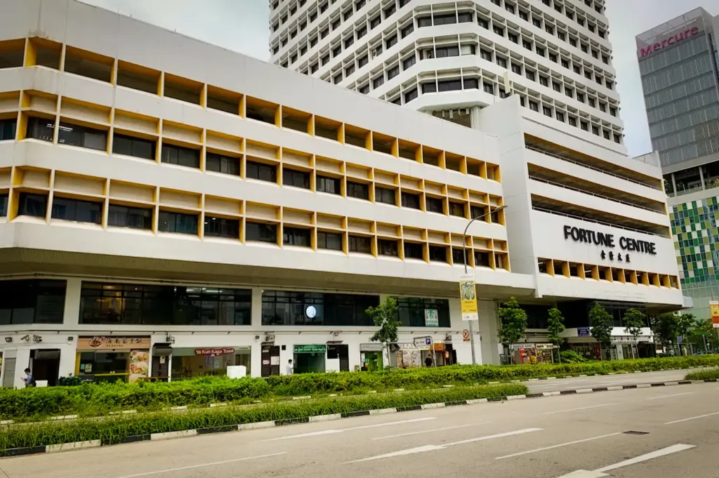 Exterior view of Fortune Centre in Singapore, showing its distinctive white and yellow facade. Street-level shops include Ya Kun Kaya Toast, set against a backdrop of urban buildings like the Mercure Hotel.
