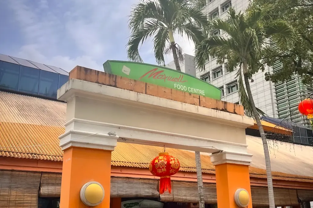 Entrance of Maxwell Food Centre in Singapore’s Chinatown district featuring the iconic hawker centre sign, tropical palm trees, traditional lantern decorations, and surrounding city skyline