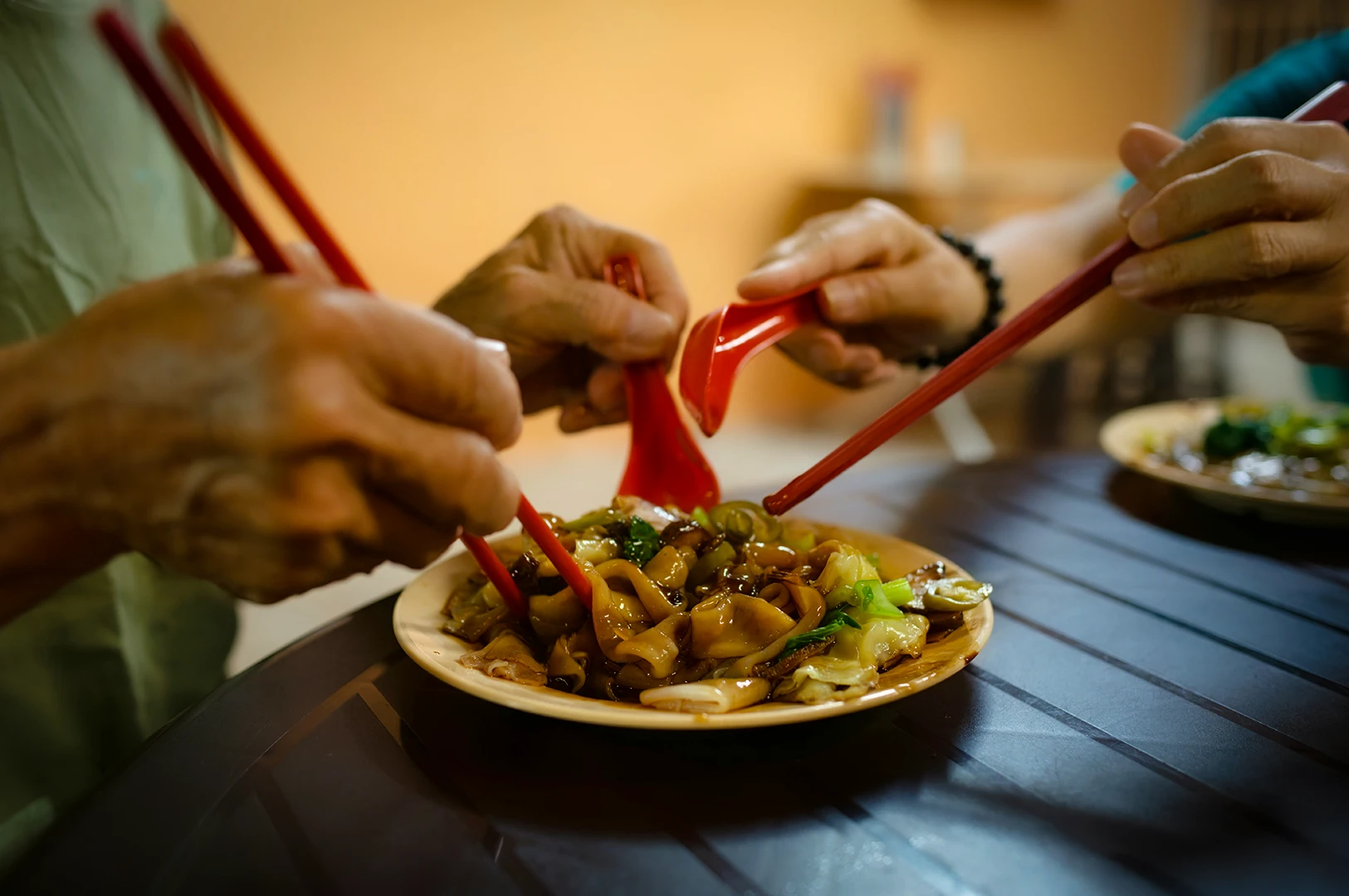 Close-up of people sharing a plate of stir-fried flat noodles using red chopsticks and spoons at a dining table.