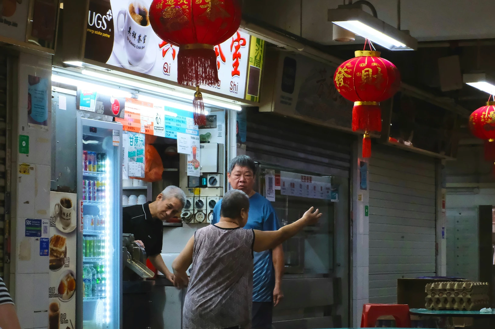 A bustling night market stall with red lanterns above. A vendor leans forward, handing an item to a customer, as another person stands nearby, creating a lively atmosphere.