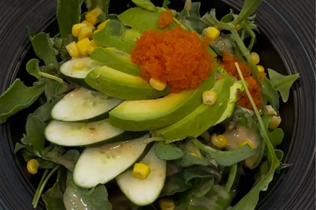 A close-up of a fresh salad served in a black bowl, topped with sliced avocado, cucumber rounds, sweet corn, leafy greens, and bright orange fish roe. A light creamy dressing is drizzled over the ingredients, creating a colorful and vibrant presentation.