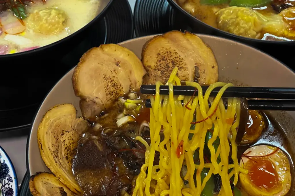 A close-up of a bowl of Japanese ramen with black chopsticks lifting bright yellow noodles from a rich dark broth. The bowl is topped with several slices of seared pork chashu, a soft-boiled egg with a jammy yolk, mushrooms, and green vegetables, while two additional ramen bowls are visible blurred in the background.