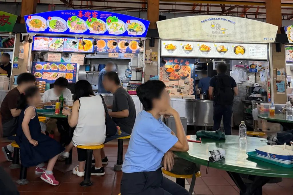 Interior of Maxwell Food Centre in Singapore featuring busy hawker stalls, communal seating tables, and local diners enjoying meals under industrial ceiling fans