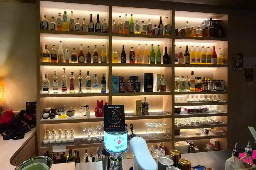 Warmly lit bar interior at UME SAN 100 in Singapore featuring shelves of Japanese spirits, sake bottles, and cocktail ingredients behind a modern counter setup