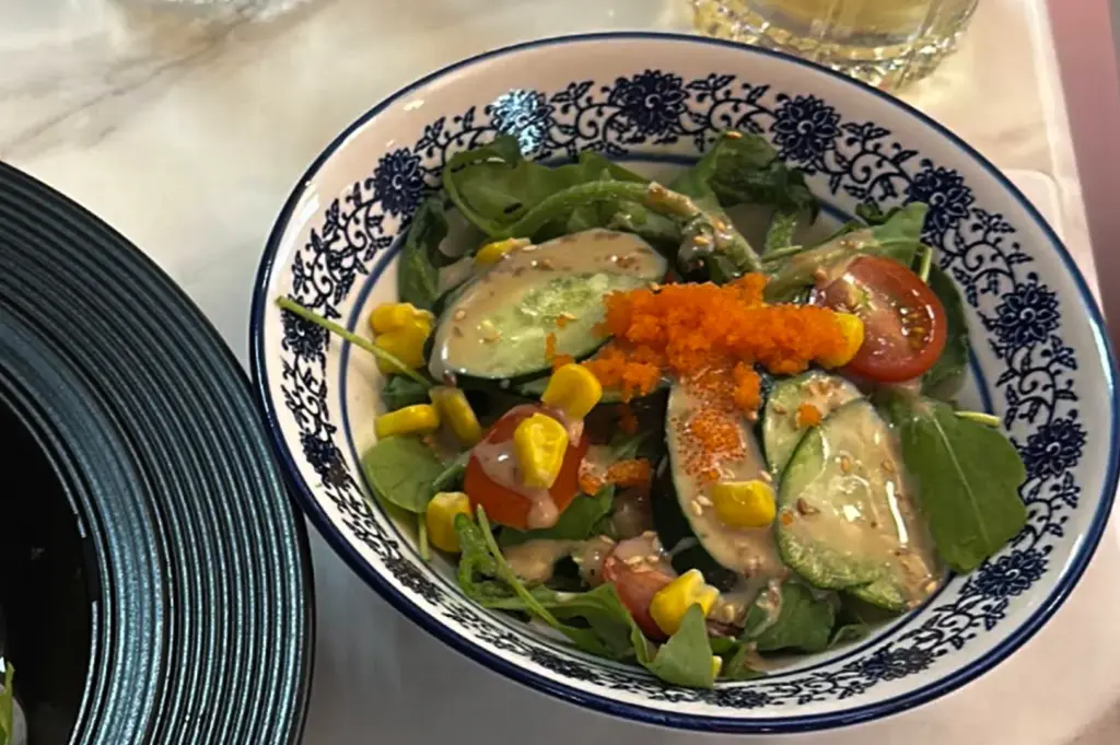 A fresh salad served in a white bowl with blue floral patterns, featuring leafy greens, cucumber slices, cherry tomatoes, sweet corn, and bright orange fish roe. The salad is lightly drizzled with creamy dressing and topped with sesame seeds, placed on a marble tabletop beside stacked black plates.