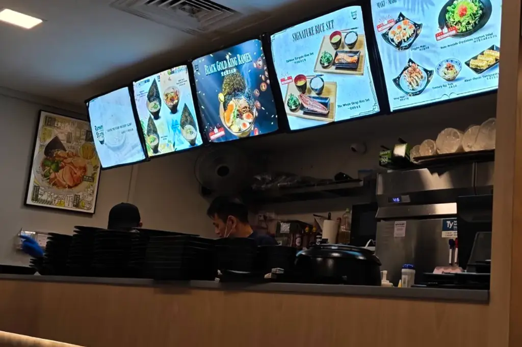 A view of a ramen restaurant kitchen counter with illuminated digital menu boards displaying ramen bowls, rice sets, and side dishes above the preparation area. Staff members work behind stacks of black bowls and kitchen equipment, while warm lighting creates a cozy casual dining atmosphere.