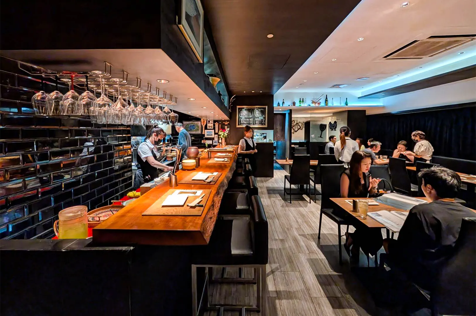 A wide, eye-level shot captures the sleek and modern interior of a bustling Japanese restaurant, featuring a long, live-edge wooden bar counter on the left where staff members in masks are busy preparing orders. Rows of polished wine glasses hang neatly from a rack above the bar, which is backed by a wall of glossy black subway tiles. To the right, several diners are seated at dark wooden tables with black high-backed chairs, some looking over menus while others enjoy their meals under soft, recessed ceiling lighting. The space is tied together by light grey wood-plank flooring and a clean, minimalist aesthetic that creates a sophisticated yet welcoming dining atmosphere.