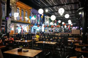 A slightly low-angle, medium-wide shot captures a lively, semi-outdoor dining area at night, representing the file "1-Beyond the Usual 5 Hidden Cuppage Plaza Food.webp". In the foreground, numerous empty wooden tables and black chairs fill a restaurant space under a high, exposed industrial ceiling, illuminated by large, glowing white globe pendant lamps and strings of colorful pennant banners. Low, black partitions patterned with hand-cutout shapes separate sections of the dining area where a few small groups of patrons are seated. In the brightly lit background, a row of historic, multi-story shophouses painted in vibrant hues of yellow, purple, blue, and green lines the pedestrian street, with their illuminated storefronts and decorative red lanterns adding a traditional charm to the festive, urban nightlife atmosphere.