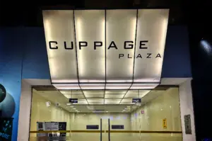 A straight-on, eye-level shot captures the modern entrance of a commercial building at night, representing the file "1-Cuppage Plaza Singapore A Quiet Food Landmark.webp". The focus is on a large, backlit sign that projects forward above a set of double glass doors, boldly displaying the text "CUPPAGE PLAZA" in a minimalist, black sans-serif font. The glowing panels of the sign extend inward to form a bright, grid-patterned ceiling over the entrance vestibule. Through the transparent glass doors, a brightly lit, empty lobby with light-colored walls and informational signs can be seen, creating a clean, structured, and welcoming urban facade.