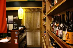 An eye-level, medium shot captures the intimate and traditional entryway of a Japanese dining establishment, representing the file "1-Keria Japanese Restaurant.webp". Dominating the right foreground is a tall wooden display shelf densely lined with various bottles of sake and spirits, many bearing individual white tags around their necks. To the left, a small wooden reception counter features credit card acceptance stickers, a telephone, and a charming maneki-neko (lucky cat) "welcome" sticker. Hanging above the counter is a glowing yellow paper lantern with Japanese characters, casting a warm ambiance over a doorway in the center background that is partially obscured by a traditional hanging beaded or bamboo-style string curtain (noren), inviting guests into a cozy and cultural space.