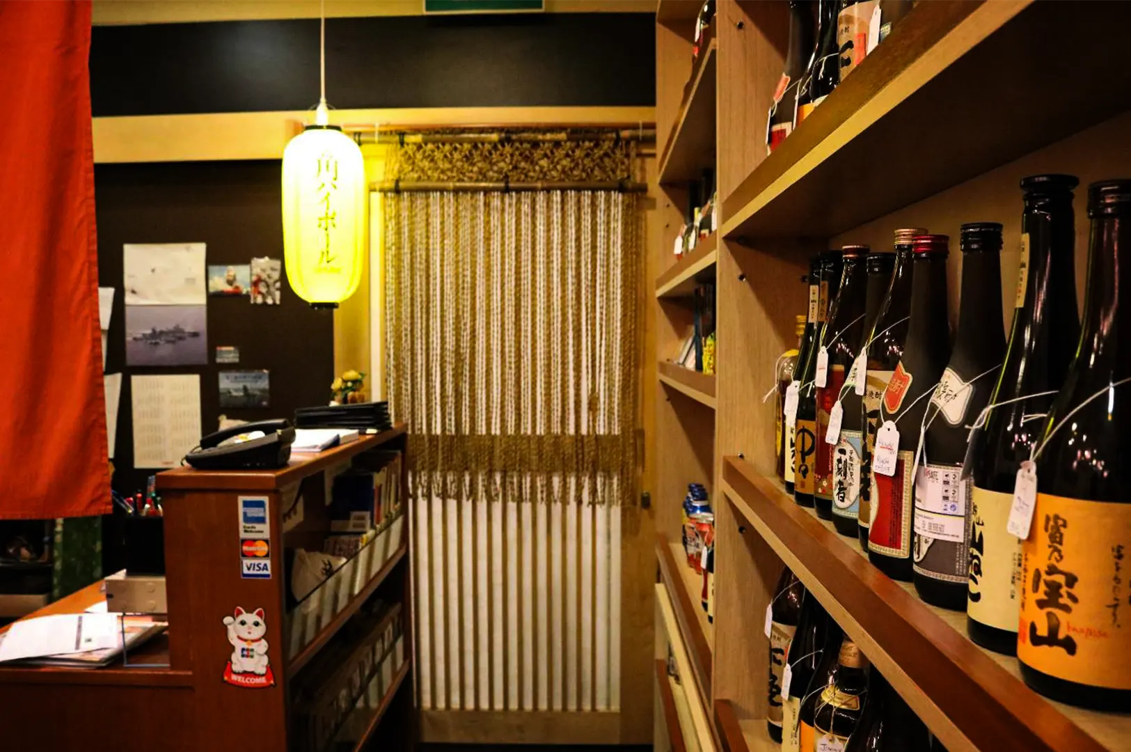 An eye-level, medium shot captures the intimate and traditional entryway of a Japanese dining establishment, representing the file "1-Keria Japanese Restaurant.webp". Dominating the right foreground is a tall wooden display shelf densely lined with various bottles of sake and spirits, many bearing individual white tags around their necks. To the left, a small wooden reception counter features credit card acceptance stickers, a telephone, and a charming maneki-neko (lucky cat) "welcome" sticker. Hanging above the counter is a glowing yellow paper lantern with Japanese characters, casting a warm ambiance over a doorway in the center background that is partially obscured by a traditional hanging beaded or bamboo-style string curtain (noren), inviting guests into a cozy and cultural space.