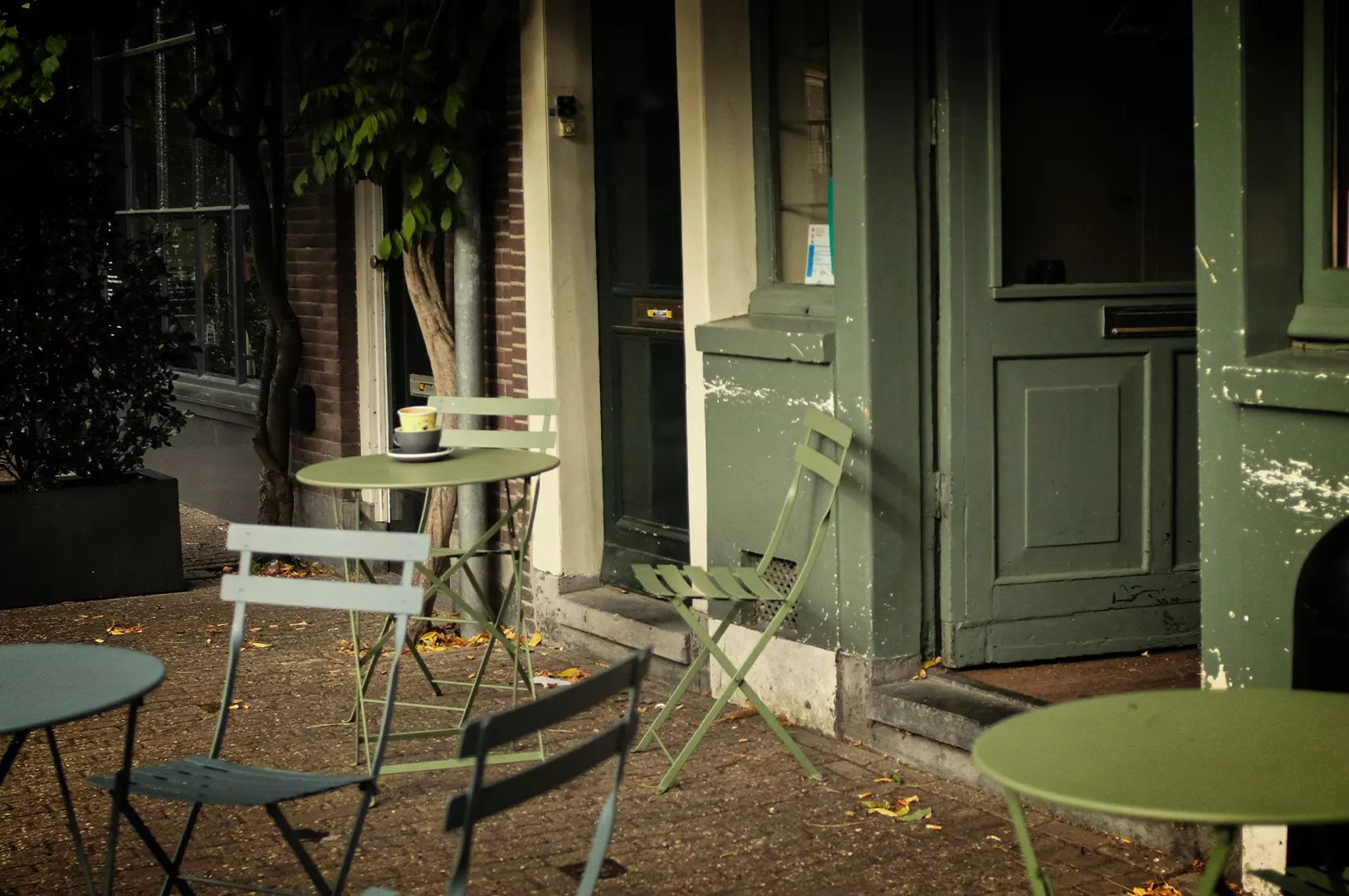 An eye-level, medium shot captures a quiet and empty sidewalk cafe setup along a brick-paved street, representing the file "1-Ten Minutes More.webp". In the center, a light sage-green metal folding table is set with two stacked ceramic coffee cups on saucers, flanked by matching metal folding chairs, while another lone green chair leans slightly against the olive-green wooden storefront wall. The background features a dark glass door and a large window pane belonging to the shop, alongside a slender tree with lush green leaves growing close to the brick facade. In the foreground, parts of two other round bistro tables and muted green chairs frame the scene, creating a serene, still-life atmosphere of a European-style street before the day's rush.