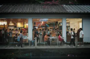 An eye-level, wide-angle shot captures a lively, brightly lit storefront bustling with people along a stone walkway, representing the file "1-The Place Everyone Walks.jpg". The single-story building features a dark, corrugated metal roof lined with small rectangular lights, contrasted by a white exterior wall with three large glass windows that showcase a vibrant interior filled with art, merchandise, and a crowd of shoppers. Outside on the grey stone-paved path, pedestrians walk by while a few individuals sit on simple chairs and stools next to small white plastic tables. In the foreground, a calm body of water with sparse lily pads perfectly mirrors the glowing storefront window displays, the silhouettes of the passersby, and a touch of purple bougainvillea flowers peeking over the roofline, creating a dynamic urban scene.