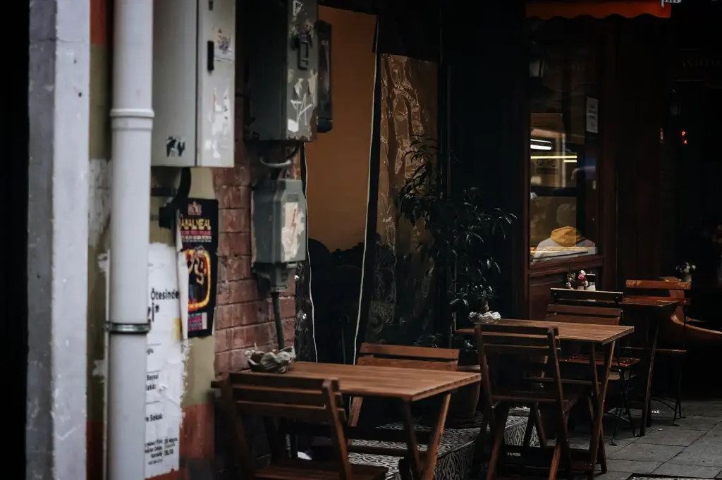 An eye-level, medium shot captures a quiet and empty outdoor seating area of a cafe nestled along a narrow urban street, representing the file "1-The Time Before the Crowd.webp". In the foreground, empty wooden slatted tables and matching folding chairs are neatly arranged on a patterned tile walkway outside a storefront. The weathered exterior features a worn brick wall mounted with industrial grey electrical utility boxes and a thick white drainpipe on the far left covered in torn flyers. The dark, shadowed background reveals a glass storefront window reflecting warm interior lighting and a hint of indoor seating, evoking a serene, atmospheric morning or late afternoon pause in a bustling city.