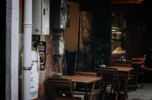 An eye-level, medium shot captures a quiet and empty outdoor seating area of a cafe nestled along a narrow urban street, representing the file "1-The Time Before the Crowd.webp". In the foreground, empty wooden slatted tables and matching folding chairs are neatly arranged on a patterned tile walkway outside a storefront. The weathered exterior features a worn brick wall mounted with industrial grey electrical utility boxes and a thick white drainpipe on the far left covered in torn flyers. The dark, shadowed background reveals a glass storefront window reflecting warm interior lighting and a hint of indoor seating, evoking a serene, atmospheric morning or late afternoon pause in a bustling city.