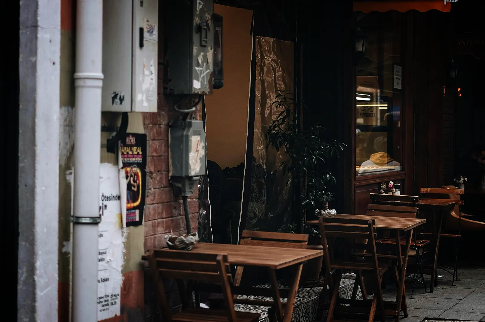 An eye-level, medium shot captures a quiet and empty outdoor seating area of a cafe nestled along a narrow urban street, representing the file "1-The Time Before the Crowd.webp". In the foreground, empty wooden slatted tables and matching folding chairs are neatly arranged on a patterned tile walkway outside a storefront. The weathered exterior features a worn brick wall mounted with industrial grey electrical utility boxes and a thick white drainpipe on the far left covered in torn flyers. The dark, shadowed background reveals a glass storefront window reflecting warm interior lighting and a hint of indoor seating, evoking a serene, atmospheric morning or late afternoon pause in a bustling city.