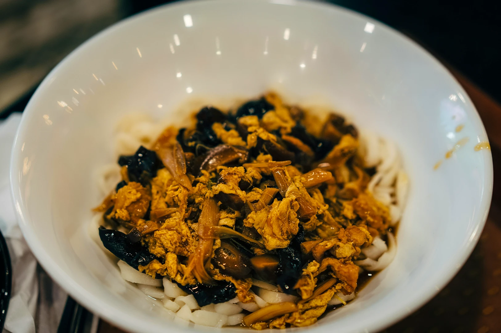 Bowl of noodles topped with braised mushrooms, shredded egg, and savory soy-based sauce in a white bowl.