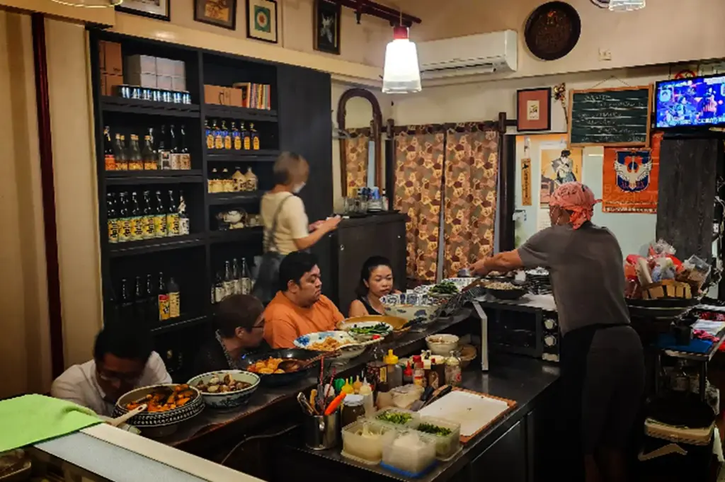 An eye-level, medium shot captures the cozy and authentic atmosphere inside a small Japanese izakaya, representing the file "2-Beyond the Usual 5 Hidden Cuppage Plaza Food.webp". A group of customers sits closely along a narrow L-shaped counter, where large ceramic bowls filled with various prepared dishes are displayed for selection. On the right, a chef wearing a pink bandana and a grey t-shirt stands behind the counter preparing food near a cooking station, while a female staff member on the left manages a tall, black shelving unit stocked with numerous bottles of sake and condiments. The background is filled with traditional decorations, including framed art, patterned curtains, a chalkboard menu, and a small television playing in the corner, contributing to the intimate, bustling dining experience.