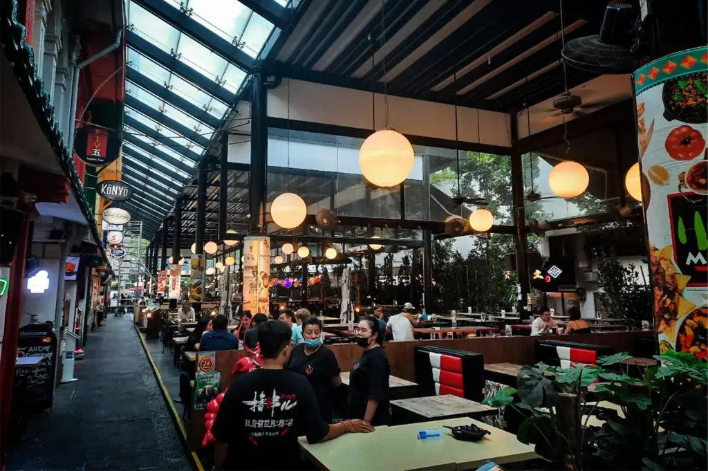 An eye-level, medium-wide shot captures a bustling, semi-outdoor dining alley flanked by restaurants, representing the file "2-Cuppage Plaza Singapore A Quiet Food Landmark.webp". On the right, patrons sit at wooden tables and red-and-white cushioned booths under a high black pavilion roof, which is illuminated by a row of large, glowing spherical pendant lamps. In the foreground, waitstaff in black uniforms converse near a service counter, while a narrow pedestrian walkway on the left stretches into the distance, lined with restaurant storefronts, hanging round signage, and a glass-and-steel awning that lets in natural daylight.