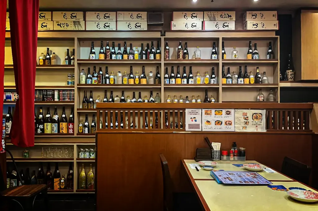 A cozy Japanese restaurant featuring wooden shelves lined with sake and Japanese liquor bottles, traditional décor, menu displays, and dining tables in an inviting izakaya-style setting.