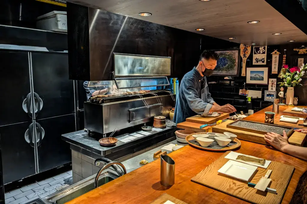 A Japanese chef prepares grilled dishes behind an open robatayaki grill at a wooden counter, while diners watch in an intimate restaurant featuring traditional Japanese décor and an authentic omakase dining experience.