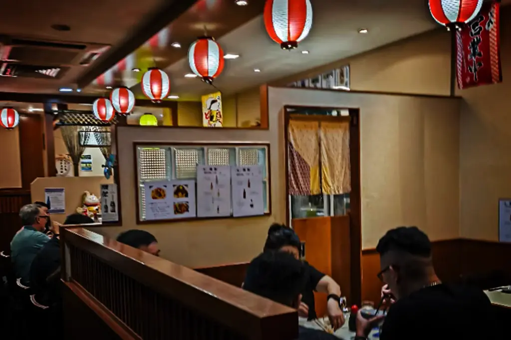 An eye-level, medium shot captures the warm and inviting interior of a Japanese restaurant, representing the file "3-Beyond the Usual 5 Hidden Cuppage Plaza Food.webp". The dining space is divided by low wooden partitions, creating semi-private booths where several patrons can be seen chatting and eating with chopsticks. Hanging from the ceiling is a long row of traditional red and white paper lanterns (chochin) that cast a soft glow across the cream-colored walls. In the background, a partition features a glass block window adorned with menu signs and a small entryway covered by a patterned noren curtain, while a maneki-neko (lucky cat) figure rests on a distant counter, enhancing the eatery's cozy, cultural charm.