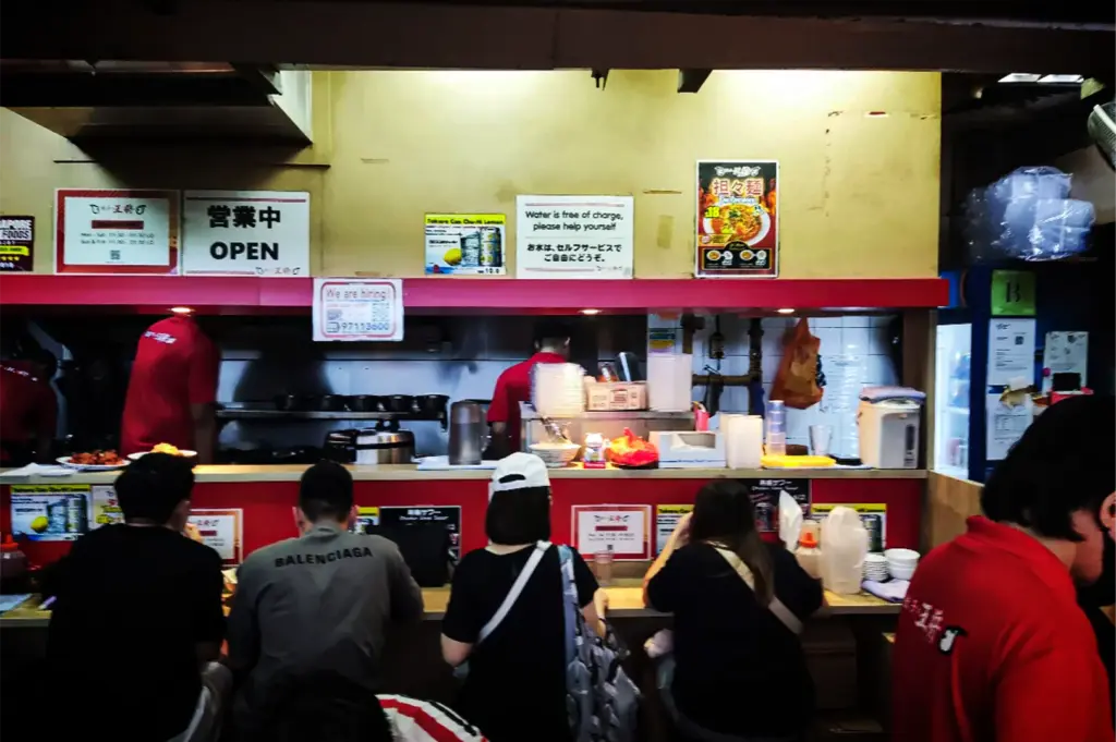 An eye-level, medium shot captures the lively and casual atmosphere of a busy ramen stall counter, representing the file "3-Cuppage Plaza Singapore A Quiet Food Landmark.webp". In the foreground, a row of patrons sits on stools along a narrow wooden counter bar, facing away from the camera while waiting for or eating their meals. Behind the counter, kitchen staff dressed in red uniforms work diligently in a compact, stainless steel food prep area filled with bowls and kitchen equipment. The counter and overhead structures are accented in bright red, while the cream-colored wall above is adorned with various functional signs, including a prominent "OPEN" notice, a "Water is free of charge" instruction in English and Japanese, and a colorful food poster, evoking the authentic charm of an indoor Asian food market stall.