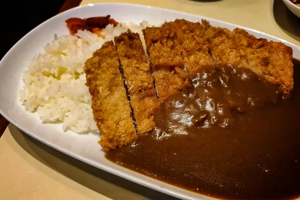 A close-up, high-angle shot presents a comforting plate of Japanese katsu curry served on a long, oval white dish, representing the file "3-Keria Japanese Restaurant.webp". A golden-brown, crispy breaded pork or chicken cutlet (katsu) is neatly sliced into vertical strips and positioned down the center of the plate. The left side is piled with a generous serving of steamed white short-grain rice, topped with a small garnish of reddish-brown pickled vegetables (fukujinzuke) at the upper corner. The right side is filled with a rich, glossy, and smooth dark brown curry sauce that partially drapes over the crispy cutlet, creating a delicious and appetizing contrast of textures.