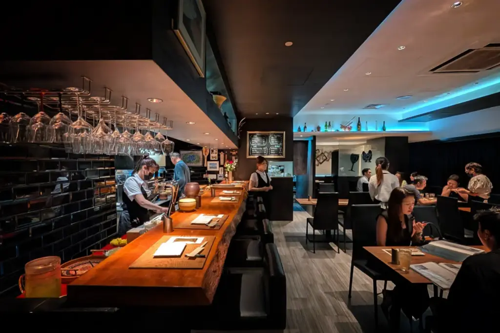 An eye-level, wide-angle shot captures the sleek and modern interior of an upscale restaurant, representing the file "4-Beyond the Usual 5 Hidden Cuppage Plaza Food.webp". On the left, a long, thick wooden counter bar with live-edge detailing serves as a focal point, flanked by black leather barstools and a black-tiled backsplash, while a vast collection of wine glasses hangs suspended upside down above the service area where staff members are working. To the right, the dining room opens up with several patrons seated at wooden tables with black chairs, engaged in conversation or looking at menus. The contemporary aesthetic is enhanced by a dark ceiling, light wood-plank flooring, and a recessed ceiling alcove on the right that radiates a cool, blue neon light, adding a sophisticated ambiance to the lively culinary venue.